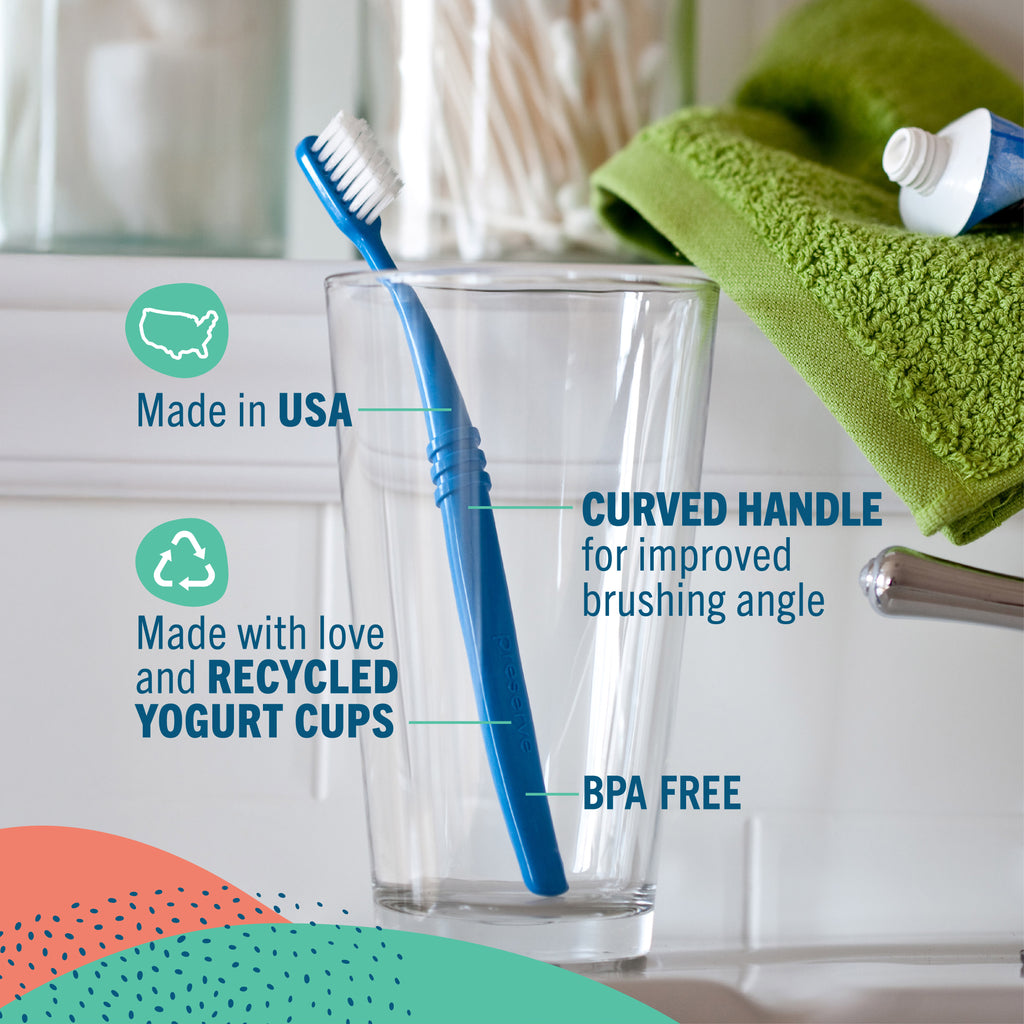 A blue Preserve toothbrush with white bristles stands in a clear glass cup on a bathroom counter, with text callouts highlighting features like 'Made in USA,' 'Curved handle for improved brushing angle,' 'Made with love and recycled yogurt cups,' and 'BPA FREE.' A green towel and toothpaste tube are blurred in the background.