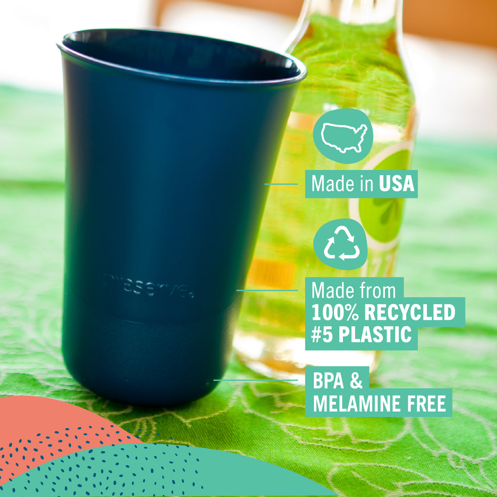 A dark navy Preserve reusable cup shown on a green patterned tablecloth beside a glass bottle, with text callouts reading Made in USA, Made from 100% Recycled #5 Plastic, and BPA & Melamine Free.