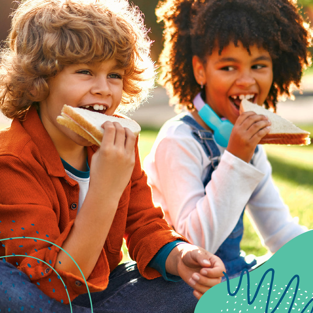 Two children eating sandwiches outdoors on a sunny day.